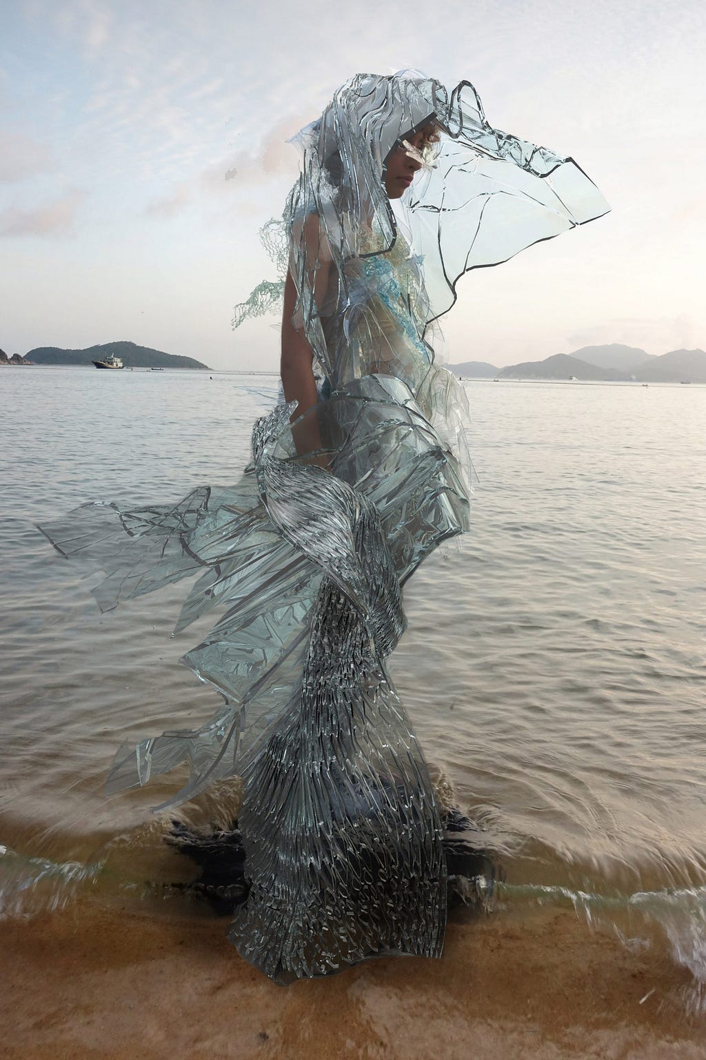 A woman is standing in a shallow part of the ocean. She wears a gown made from a light blue, slightly translucent stretches of material that flow out behind her, with the bottom skirt made from a net-like material.