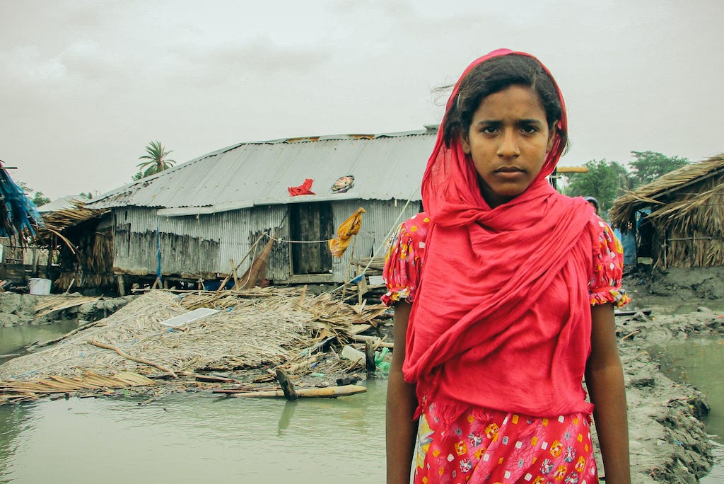 Aduri is wearing a red dress and headscarf and standing in front of her collapsed home.