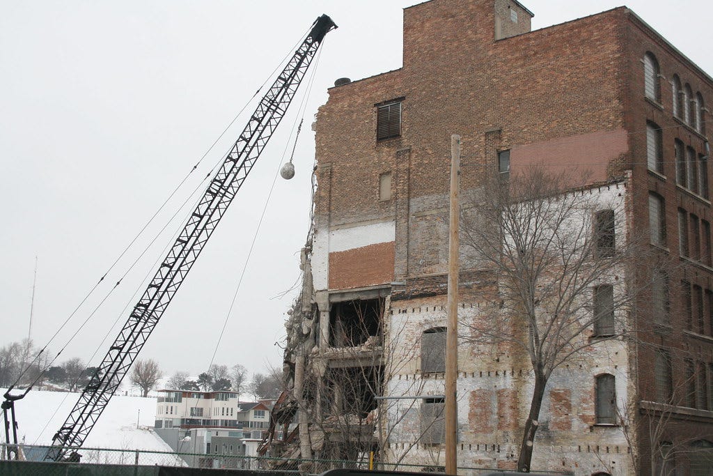 A wrecking ball on a crane swinging towards an old partially-wrecked brick building.