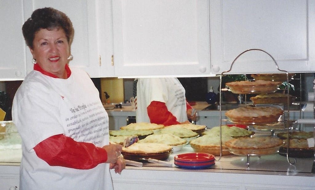 My adoptive mother, wearing a white short-sleeved T-shirt over a red long-sleeved one, smiling and standing by a counter with multiple mincemeat pies on it.