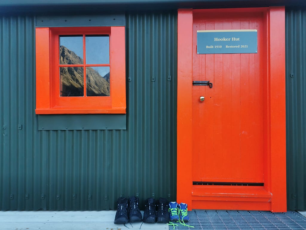 Two adults and one childs pair of boots outside the red door of the Hooker Hut. A sign reads “Hooker Hut, Built 1910, Resoted 2021”