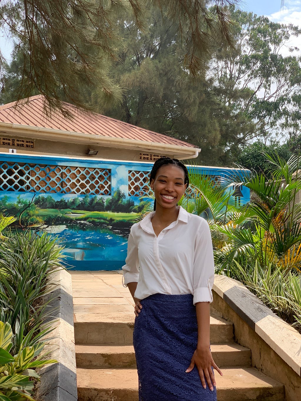 Stevie, a Black woman with her dark hair in braids pulled back, stands in a tropical setting in Uganda on a paved sidewalk in front of a couple stairs. She has one hand on her hip and the other by her side. She is wearing a cream button-up blouse and a dark blue skirt. She is looking at the camera and smiling.