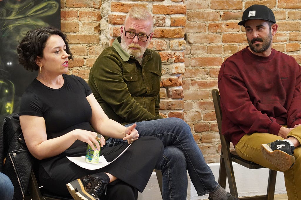 Three people sit in conversation. One woman with dark cropped hair wearing a black t-shirt speaks while two men look on listening attentively.