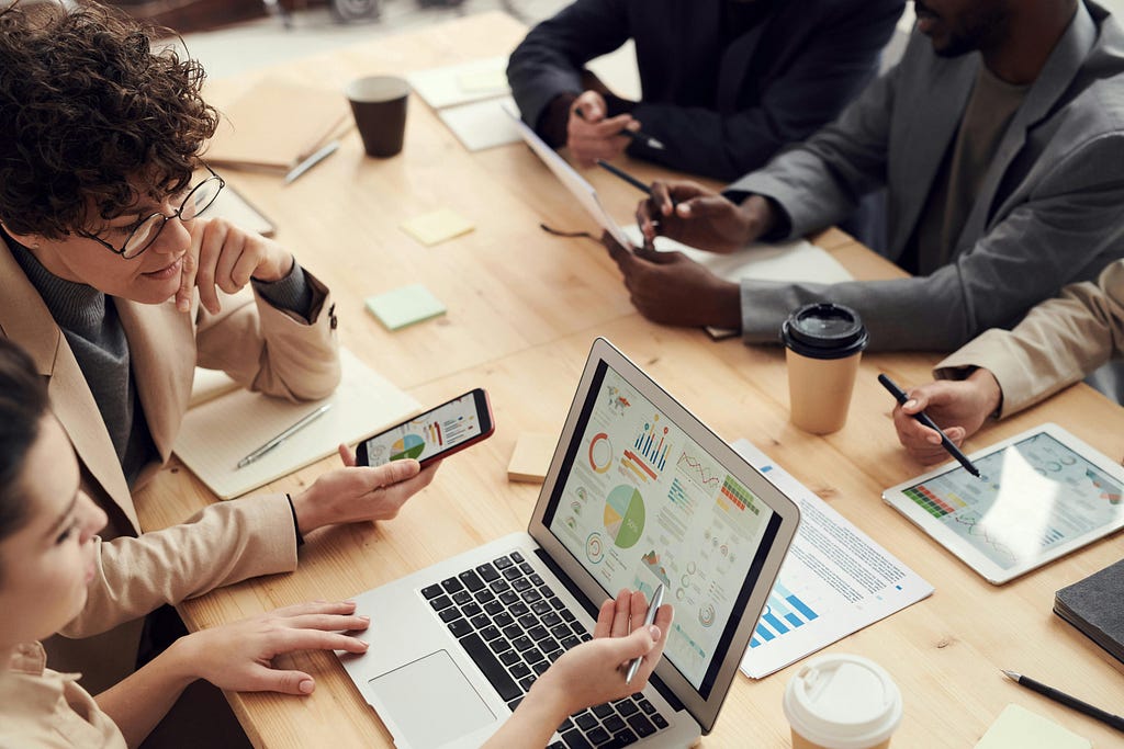 Grupo de pessoas em uma reunião ao redor de uma mesa de madeira clara. Na mesa, há diversos dispositivos tecnológicos, como laptops, tablets e smartphones, exibindo gráficos e dados. Há também papéis, canecas de café, e canetas espalhadas pela mesa.