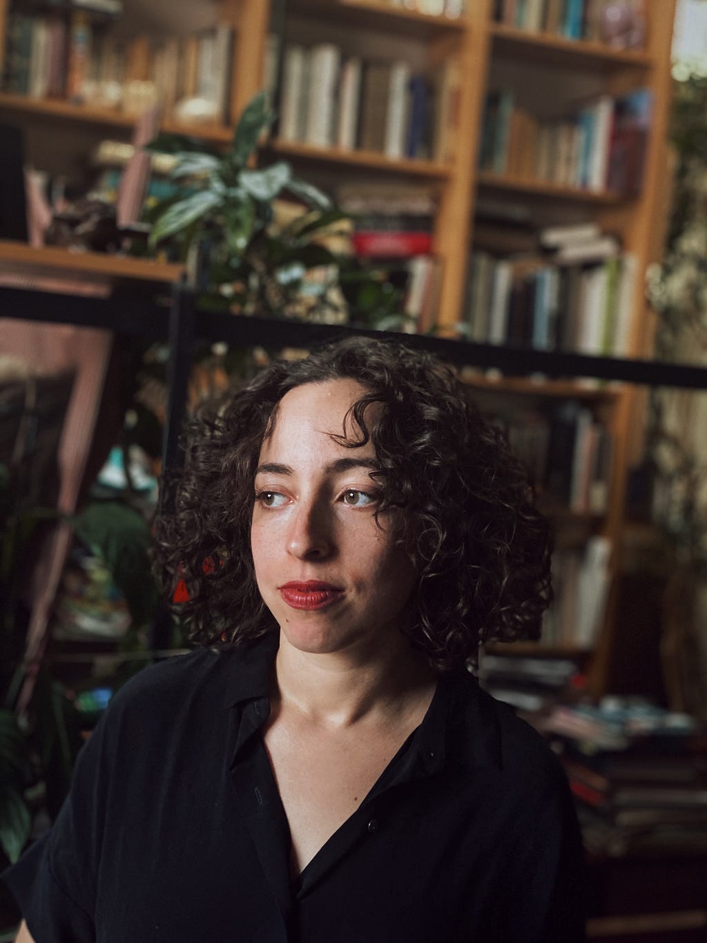 Woman with curly hair in a black shirt, thoughtful in a room full of bookshelves.