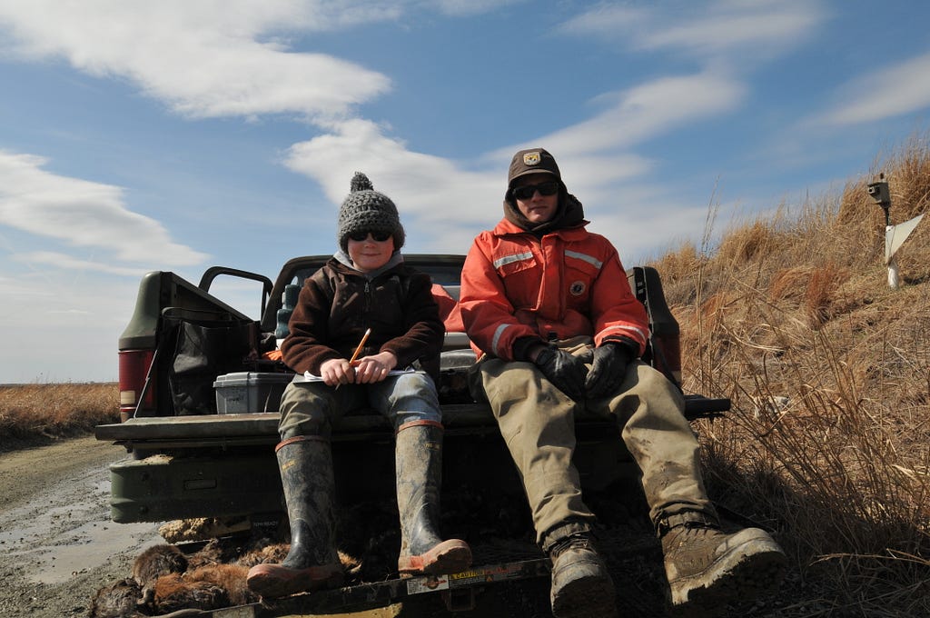 A small boy wearing rubber boots and a knit hat sits on the tailgate of a pickup truck, beside a man in wading boots, an orange coat, and a U.S. Fish and Wildlife Service cap.