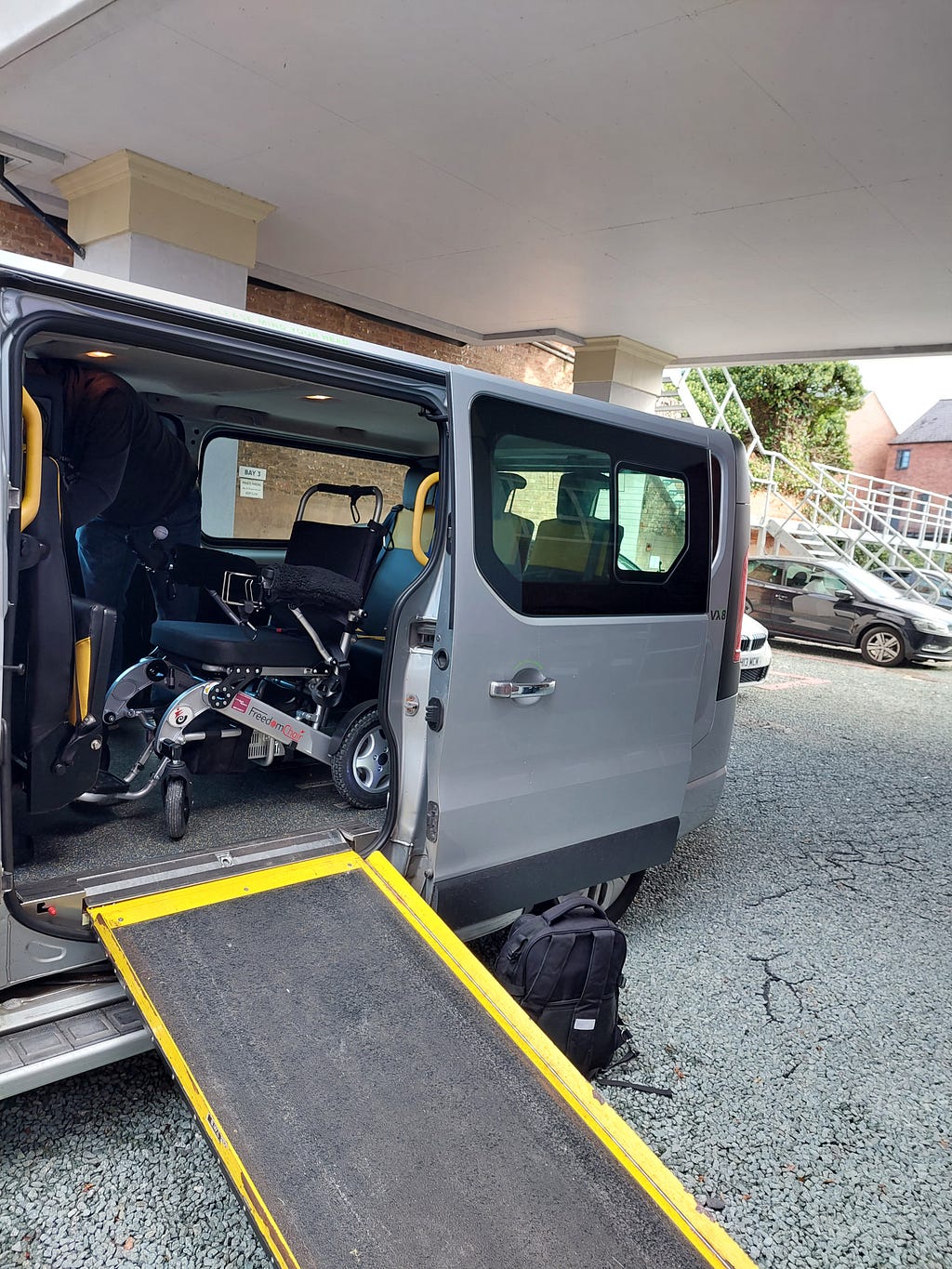 Claire’s chair secured inside one of Dell’s Taxis wheelchair accessible vehicles. The vehicle is a mini-bus-style vehicle. There is a ramp that has been slid out from the side of the vehicle. The ramp and all handles are yellow, to aid visibility.