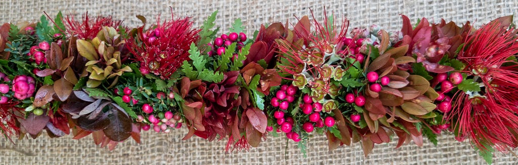 Photo of a lei made wili (twist; wind) style with red lehua (Metrosideros polymorpha) blossoms, dark red liko (M. polymorpha), pink pūkiawe (Leptecophylla tameiameiae) fruit, and green palapalai (Microlepia strigosa) fern.