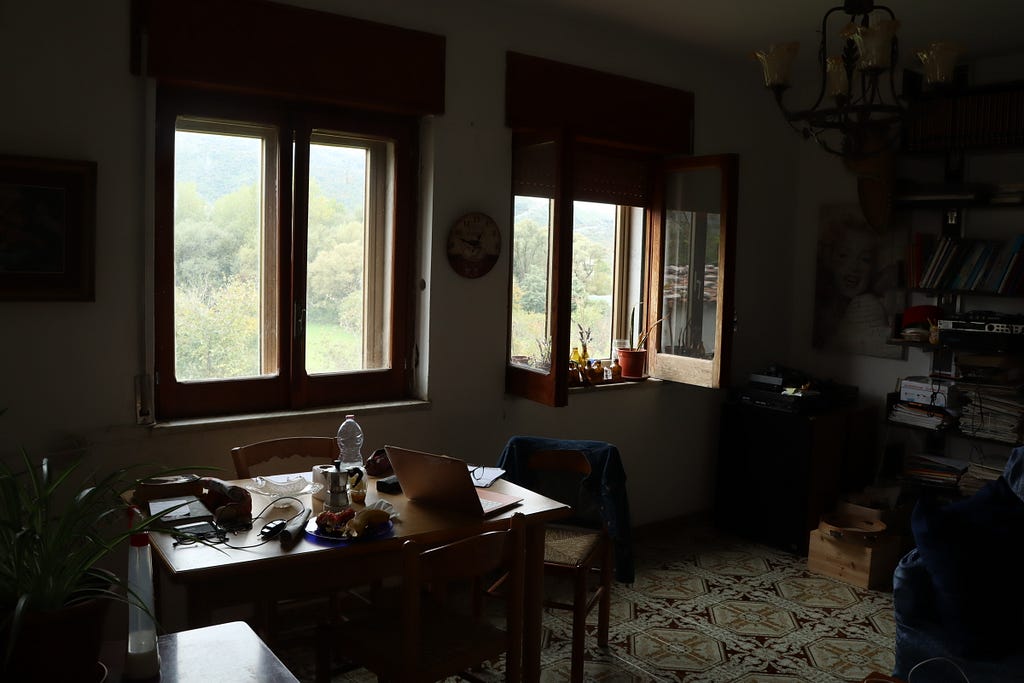 Cozy Italian farmhouse interior with open windows revealing verdant countryside fields, fruit trees, and rolling hills, offering a peaceful rural escape near Naples.