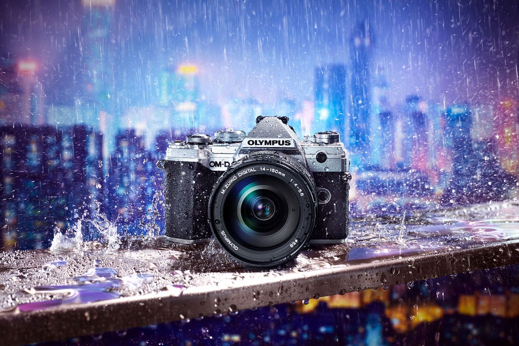 Weather sealed EM5 III camera under heavy rain, on a balcony barrier edge with lit up city sky scrapers in the background at night.