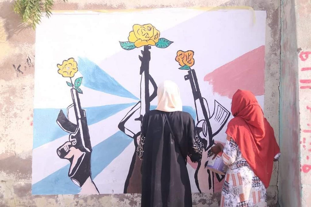 Two women with their backs to the camera gaze at a mural on an outdoor wall that shows three rifles pointed into the air with a flower coming out of each barrel.