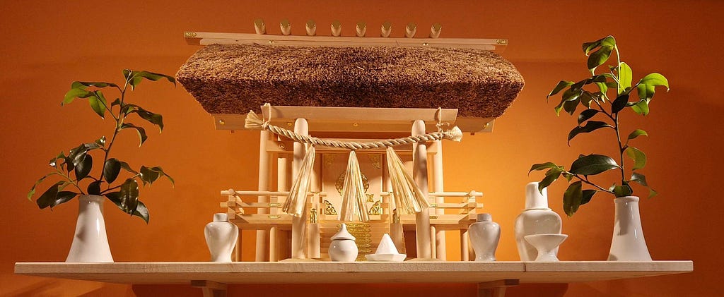 A small wooden structure with a thatched roof and miniature hemp rope spanning the entrance. Hemp tassels hang from the rope. Small white ceramic vessels stand in front of the structure. On either side is a small white ceramic vase with a branch of green leaves in it. The whole thing is on a wooden shelf in front of a peach-coloured wall.