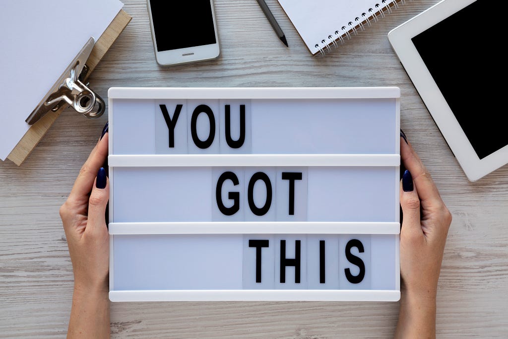 A person holds a sign that says, “You got this.” A notebook, iphone, and ipad are nearby on a gray desk.