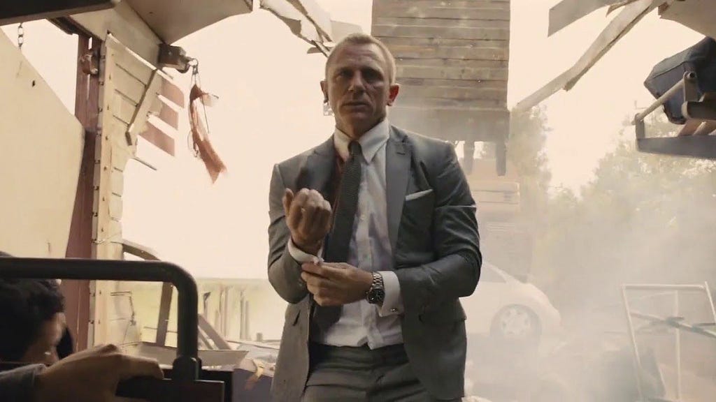 Daniel Craig a middle aged actor playing bond is standing on a train carriage with the scoop of a digger having crashed through the ceiling and absolute carnage behind him. He is adjusting his shirts cuff.