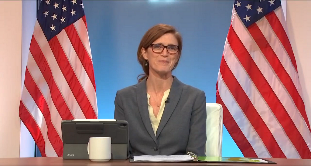 Flanked by two American flags, USAID Administrator Samantha Power sits at a table with a tablet and a three-ring binder open and a white drinking mug propped behind the tablet.