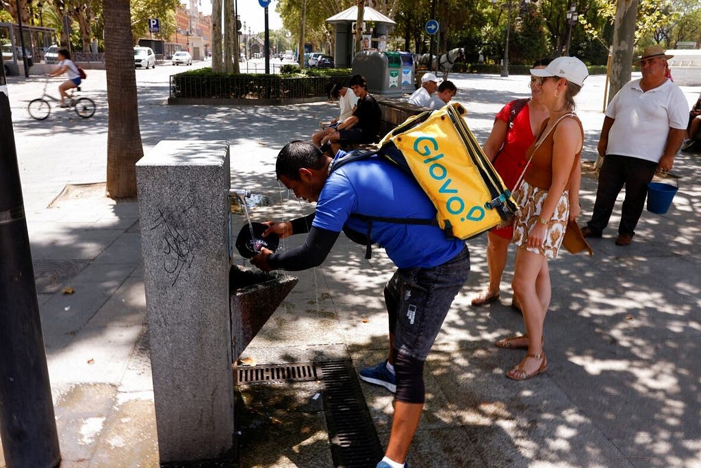 Photo taken on a public place in Seville, Spain. A delivery rider with a Glovo bag is water their hat in a public fountain to refresh themselves. People are queuing behind them to access the water fountain.
