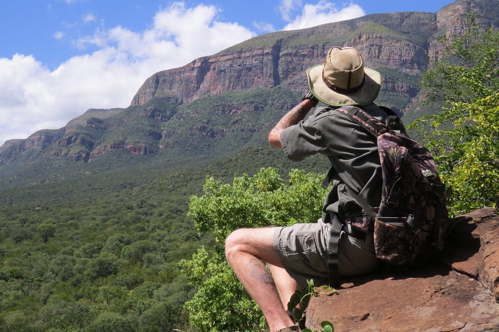 A Caucasian male wearing a trail guide uniform, sitting on a rock, looking through binoculars at a mountain range