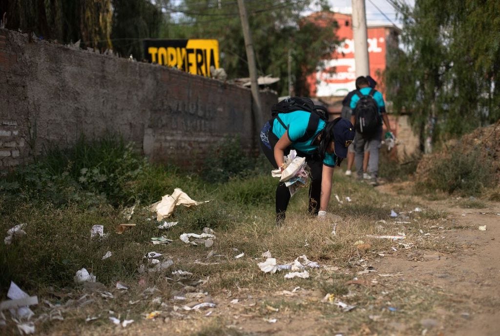In Bolivia, teenagers wearing blue t-shirts are picking up trash. There is a brick wall to one side.