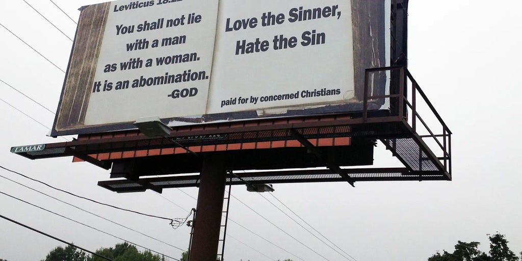 billboard with edges painted to look like an open book. On the left page, text reads “You shall not lie with a man as with a woman. It is an abomination.” — God. Right page reads “love the sinner, hate the sin. Paid for by concerned Christians.”