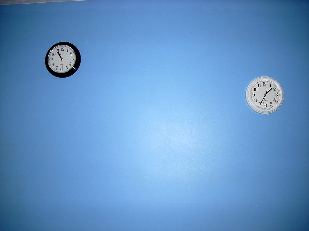 a small black clock and a small white clock mounted on an otherwise empty wall