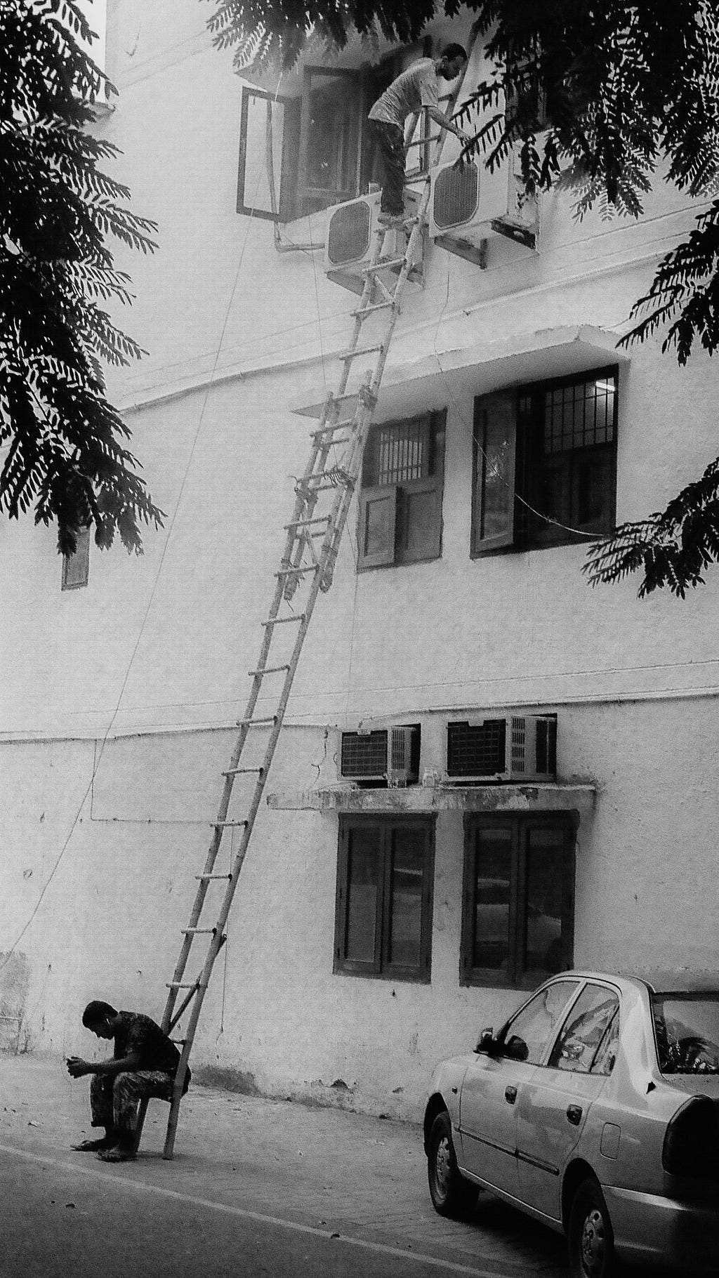 Black and white image of two men on a tall ladder one sits at the bottom while one works near the top.