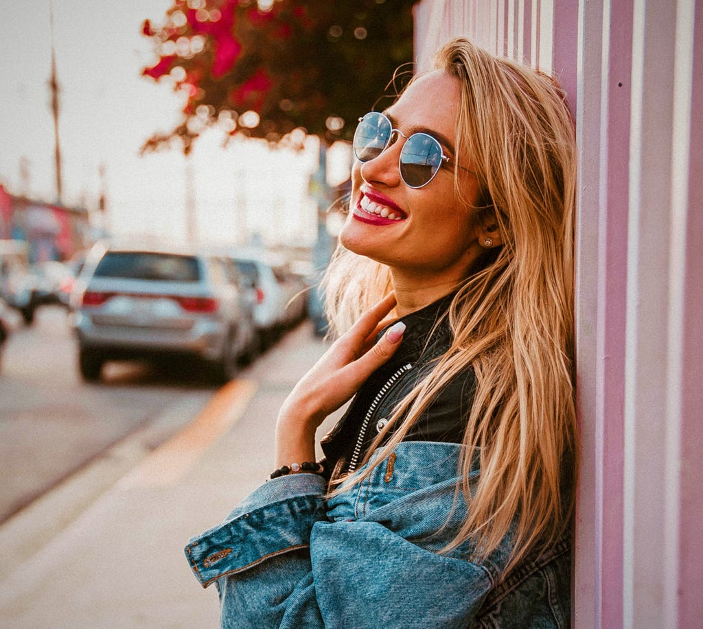 smiling girl wearing sunglasses leaning against wall with hand laying on her neck