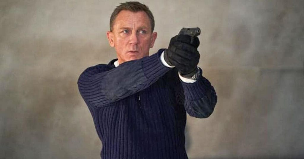 Daniel Craig , a middle aged actor playing bond holds up a handgun with a no-nonsense expression on his face. He is wearing combat gloves and a fisherman’s style hard wearing jumper.