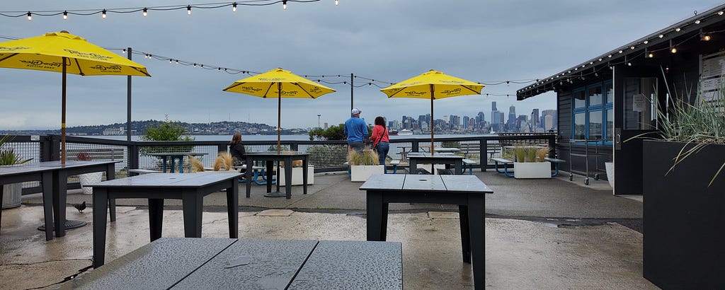 Outdoor seating at Marination with yellow umbrellas, wet tables, and a view of the Seattle skyline across the water on a rainy afternoon.