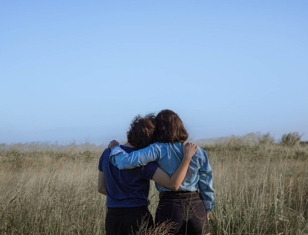 Two people stand in a paddock with their arms around each other and resting their heads together.