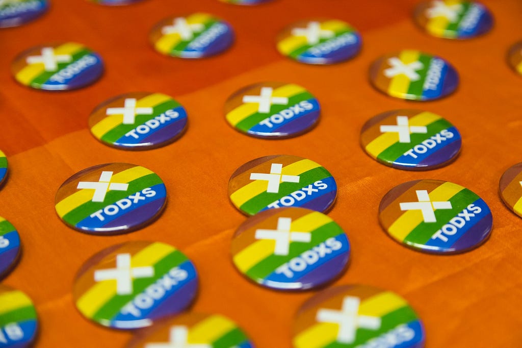 A imagem é uma foto aproximada de vários bottons com estampa de arco-íris e o logo da TODXS em branco no centro.