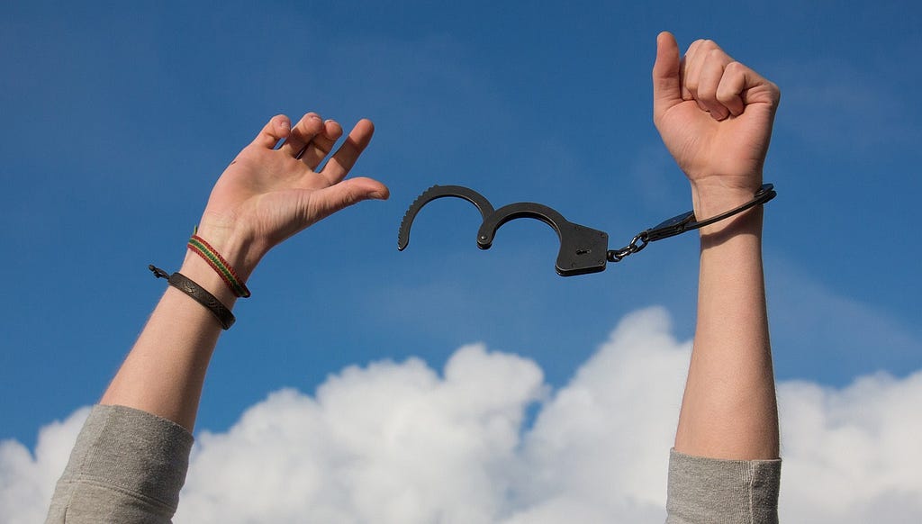 A photograph shows a person’s forearms raised up. Gray shirtsleeves are barely visible at the bottom of the image. The person has a woven or beaded multicolor bracelet and a dark colored leather bracelet on their right wrist. They have one locked handcuff on their left wrist. Attached is an open handcuff that swings in the air between their hands. A blue sky with puffy white clouds is shown in the background.