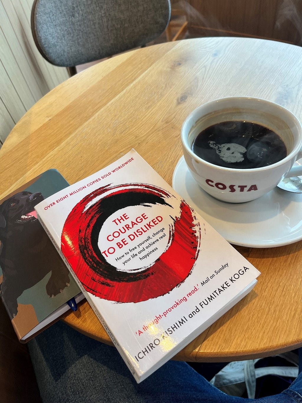 ‘The Courage to be Disliked’ book laying ontop of a notebook with a black labrador on, next to a costa mug with steaming Americano coffee in.