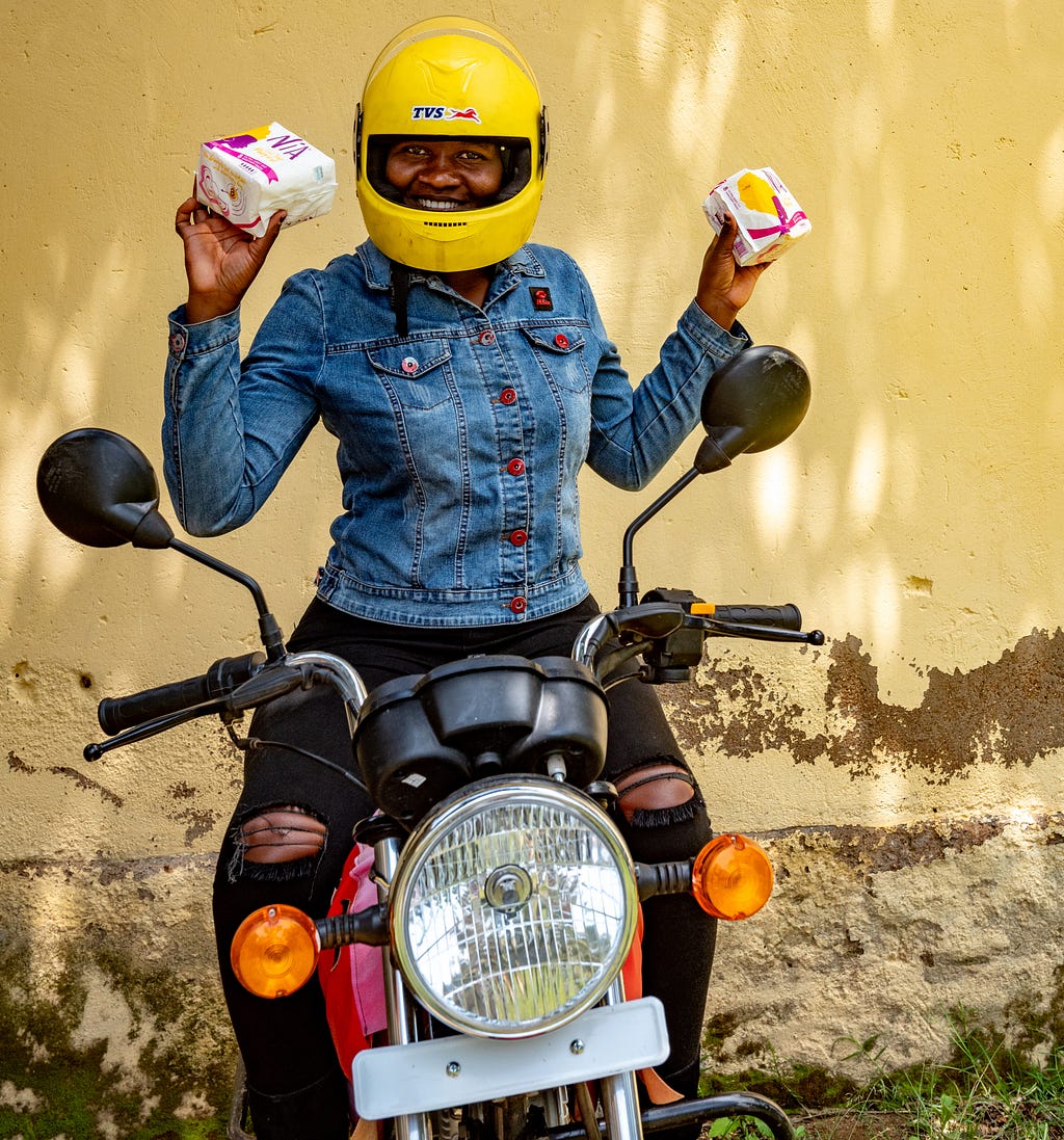 A smiling woman on a motorbike with a yellow helmet holds up menstrual pads.