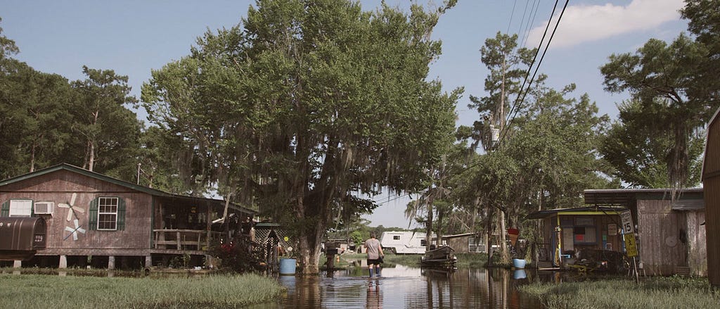 Film still from Belle River showing the back of a man in a grey t-shirt, black shorts, and rubber boots, standing underneath a tree and up to his calves in calm water, which is filling the space between two houses. The house to his left is built on stilts.