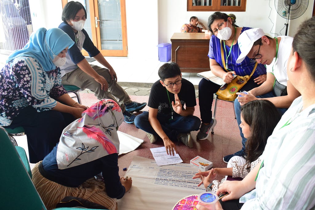 A group of eight people sat in a circle at a workshop. A man is sat on the floor explaining something, while the other seven people are listening.