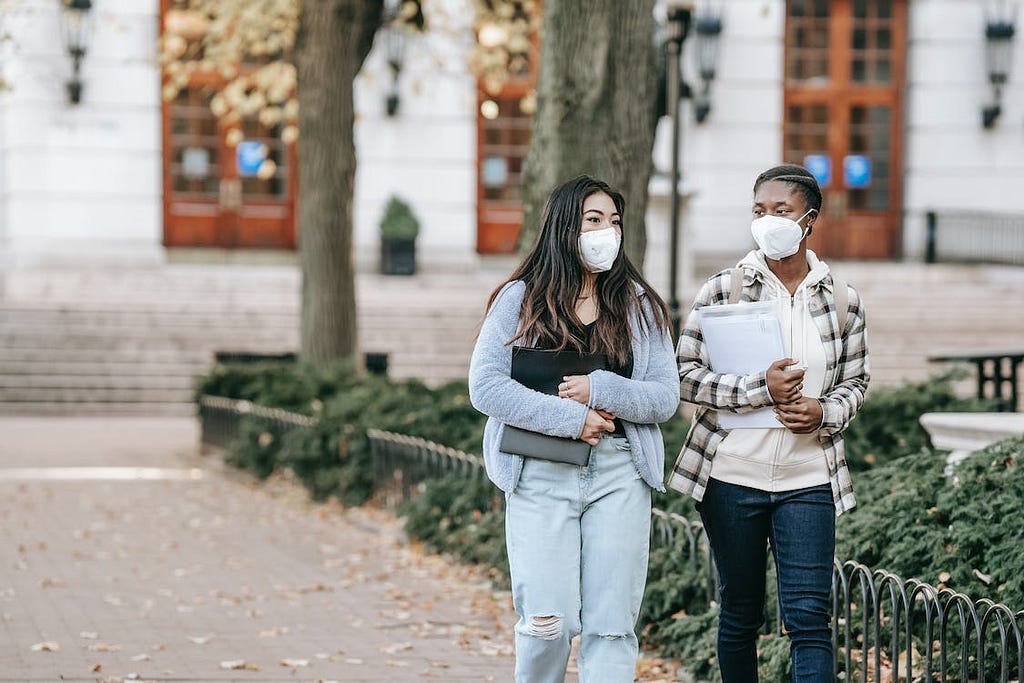 Two young women are walking outdoors wearing white KN95 masks. They are strolling down a paved walkway next to some greenery. The setting appears collegiate; they look like two students, carrying papers and wearing jeans and sweaters. They appear to be mid-conversation.