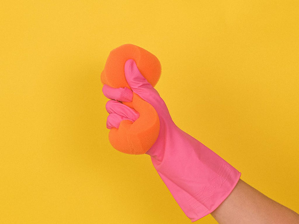 A hand wearing pink cleaning gloves holds an orange sponge against a yellow wall.