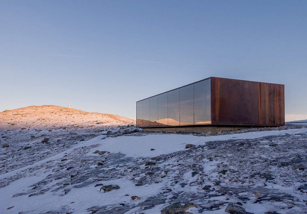 A long, rusty structure with a glass wall sits on a mountainside surrounded by snow.