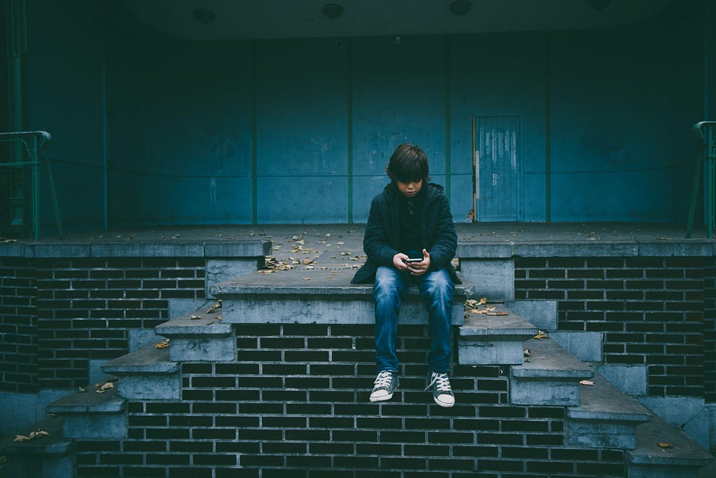 Um menino, vestindo um casaco, calça jeans e um tênis all star sentado no alto de uma escada interagindo com o seu smartphone