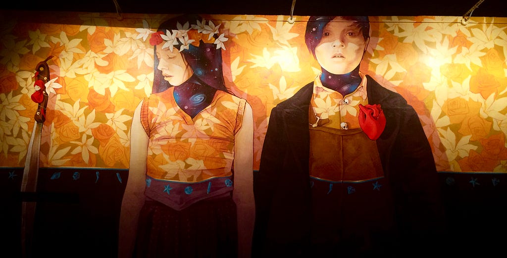 Image of a painted wall by street artist INTI of two people standing elbow to elbow one looking forward and the other to the side. Both have yellow shirts with leaves that are the same as the background. There is an anatomical red heart on the  outside the clothing of the one looking forward.