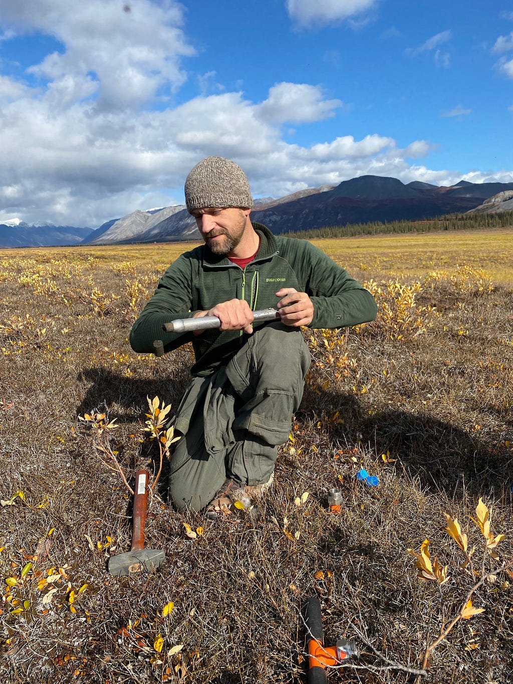 A man with a soil core sampler on an open landscape with mountains in the distance.