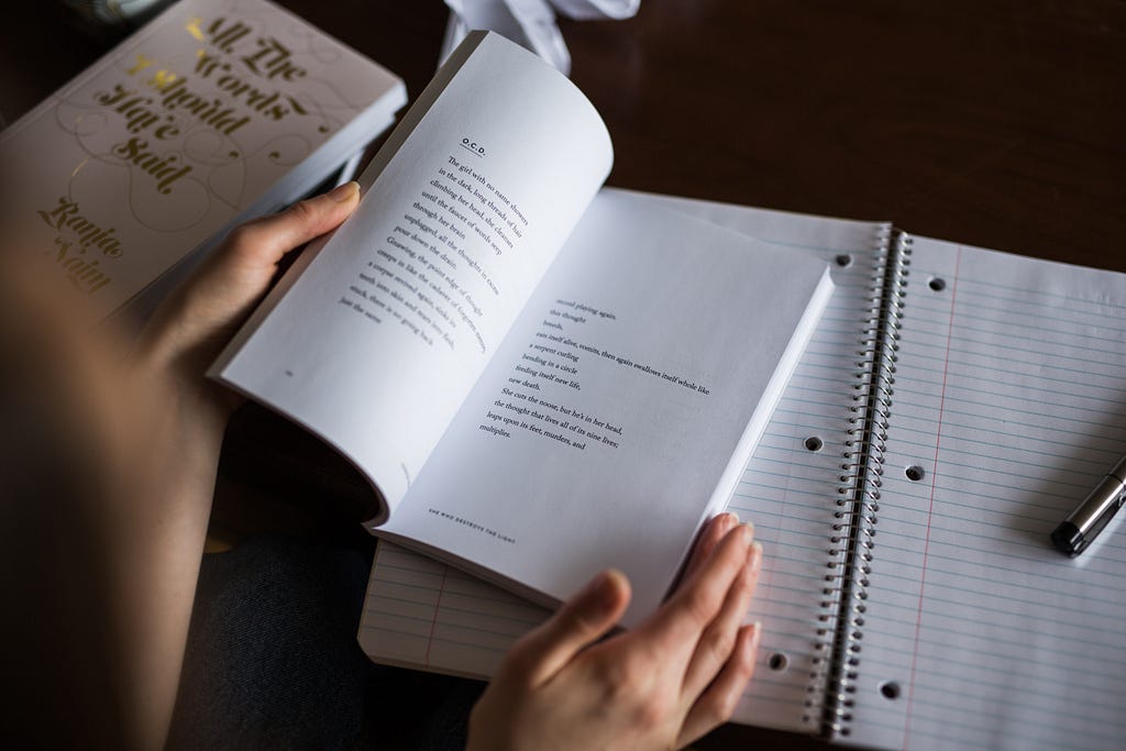 A young woman is pictured reading poetry from an open book, placed above empty notes from a large notebook.