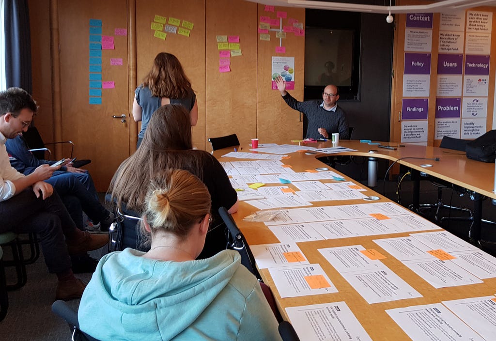 The team sit around a table with papers on as Tom Steinberg gestures to a wall of post it notes clustered together.