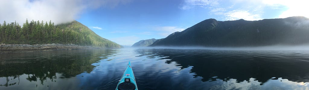 A panorama centred around the nose of a kayak, steep mountains on either side and an ocean channel with low fog in centre