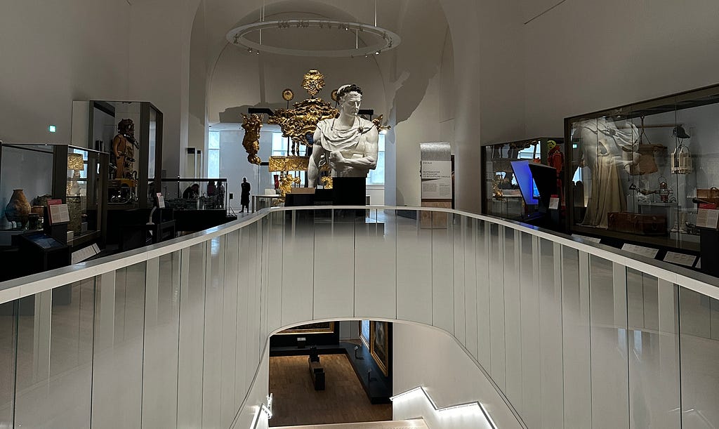 Inside the Musée de la Marine, a white marble bust stands prominently on a balcony with glass barriers. Below, a circular opening reveals a lower floor. The room is well-lit, with displays of various artifacts in glass cases. A golden dragon boat sculpture is visible in the background.
