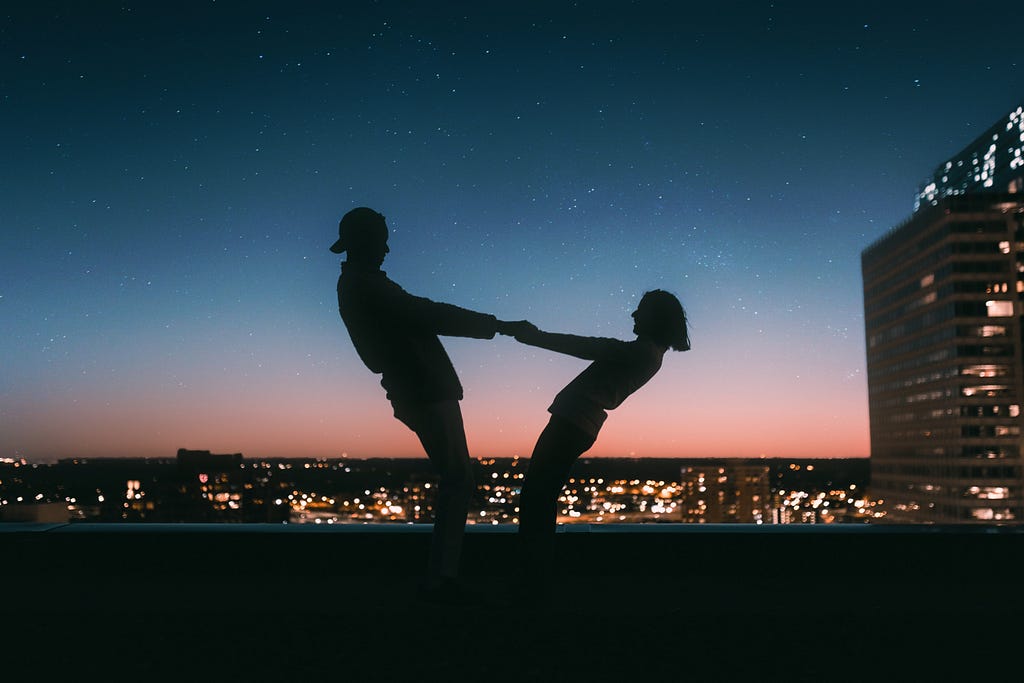 Two human silhouettes holding both hands, stretched away from each other, with the last glimpse of sunset and twinkling lights behind them.