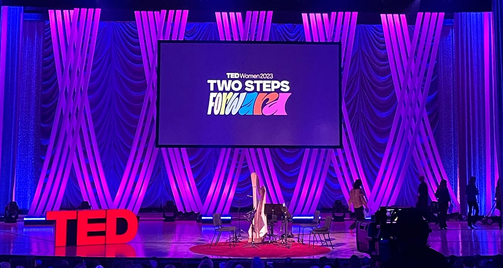 A color photograph of the TEDWomen stage, with the big red TED letters on the left, instruments set up for a band, purple-hued lighting, and a screen that reads “TEDWomen2023: Two Steps Forward”.