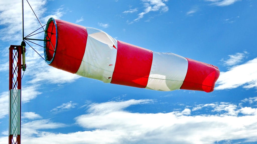 a windsock blowing in the wind. The windsock is attached to a tower and there is a partly cloudy sky in the background on a sunny day.