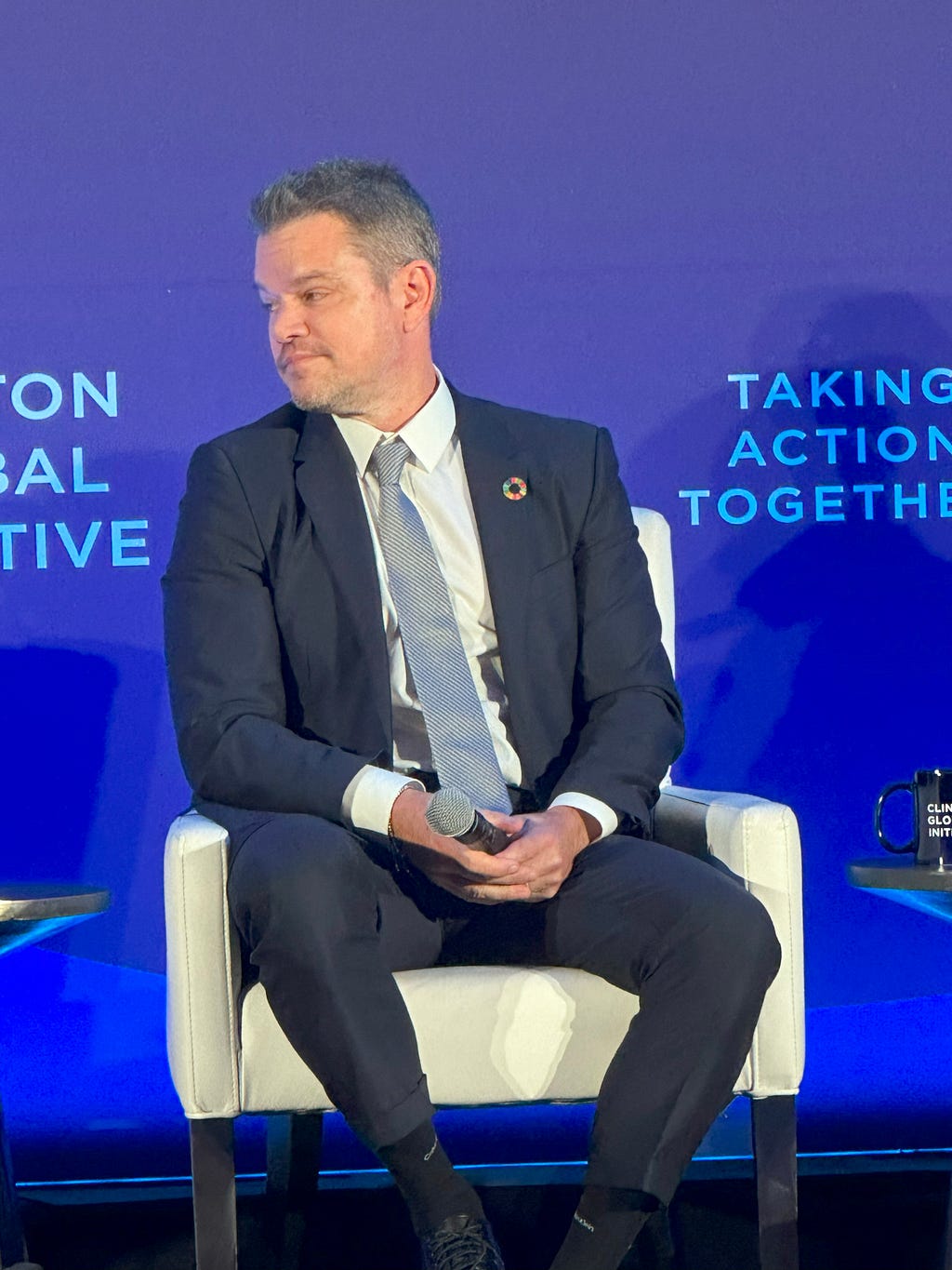 Matt Damon wearing a navy suit, white shirt and blue tie against a blue background. He is sitting in a white chair and looking to his right.