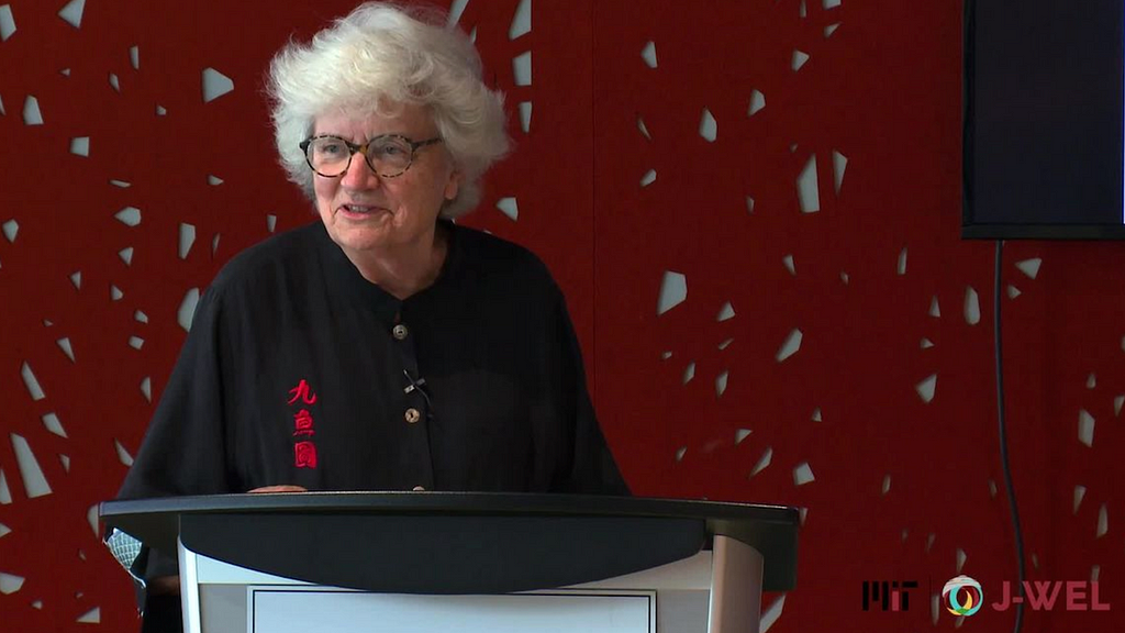 Photo of an older woman wearing glasses and a clip-on microphone, speaking behind a podium.